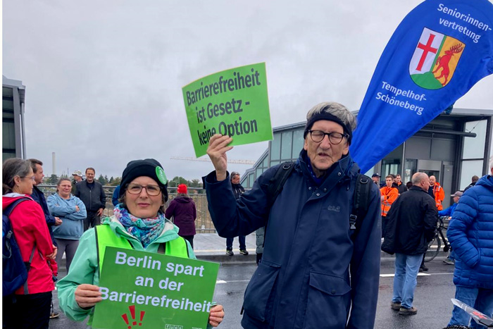 Wolfgang und Annabelle halten Schilder von FUSS e.V. zum Thema Barrierefreiheit am Eingang des S-Bahnhofs hoch
