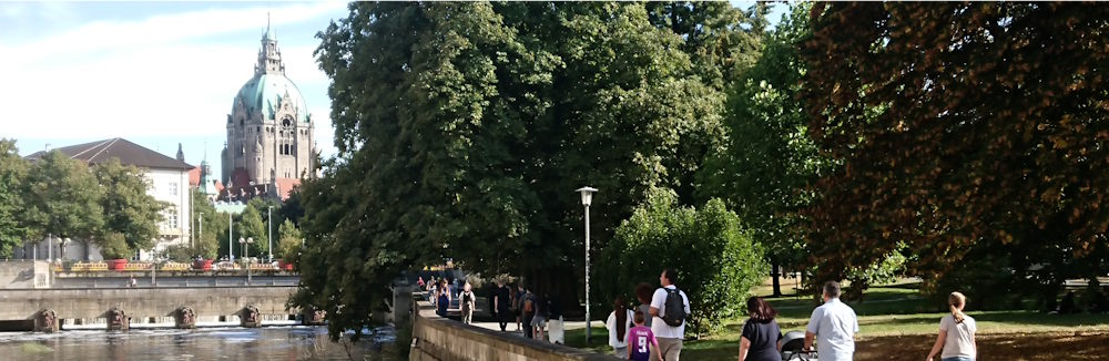 Fußgänger_innen am Leineufer in Hannover. Im Hintergrund das Neue Rathaus.