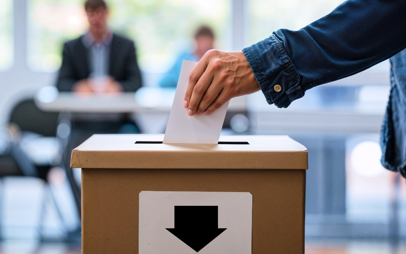 Im Vordergrund steckt jemand einen Wahlzettel in eine Wahlurne, im Hintergrund sind unscharf zwei Personen an einem Tisch sitzend zu sehen