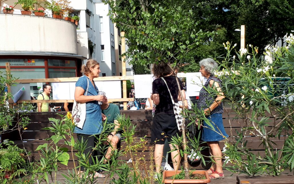 Mehrere Menschen stehen auf einem sonnigen, durch Topfpflanzen begrünten Platz und unterhalten sich.