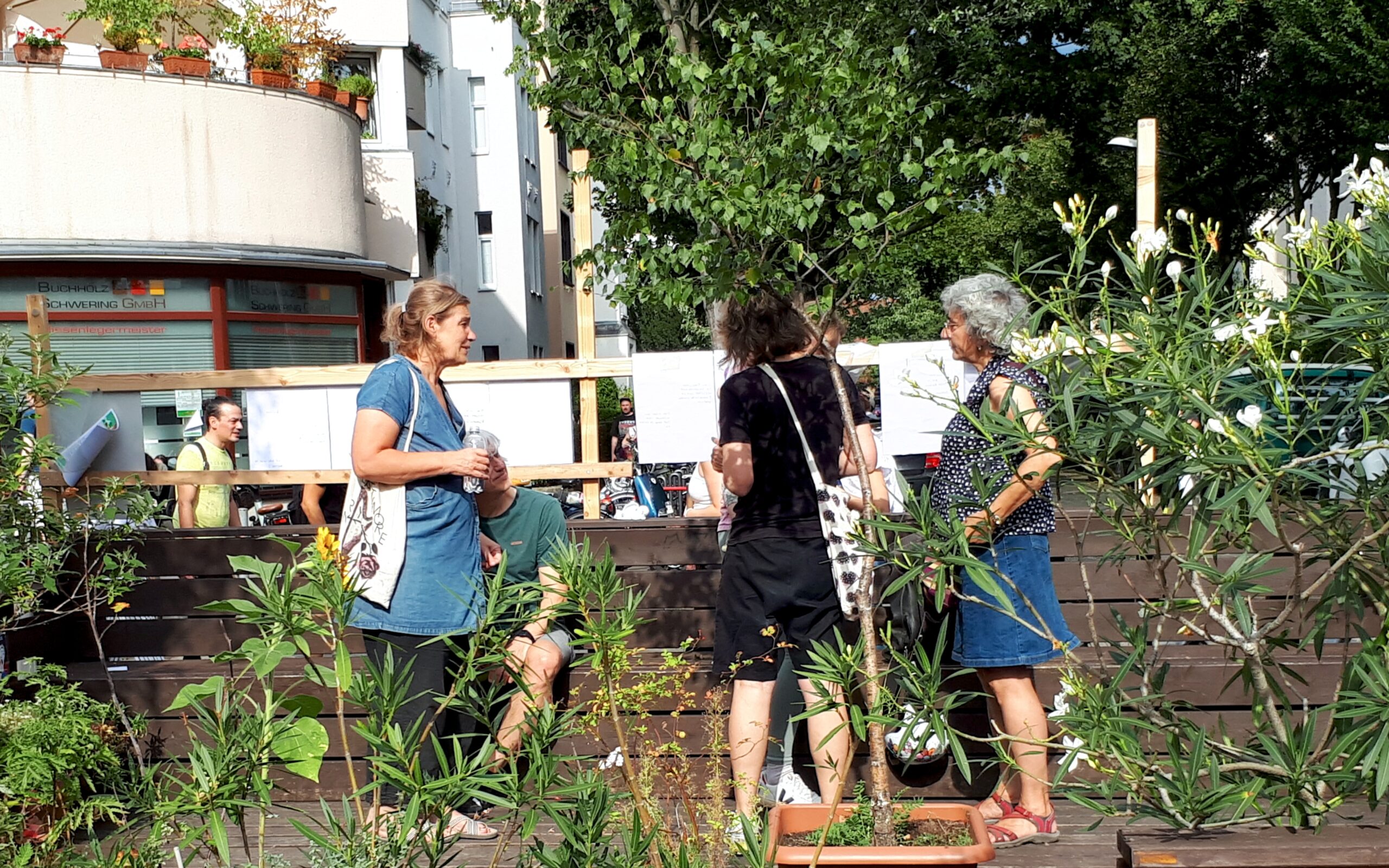 Mehrere Menschen stehen auf einem sonnigen, durch Topfpflanzen begrünten Platz und unterhalten sich.