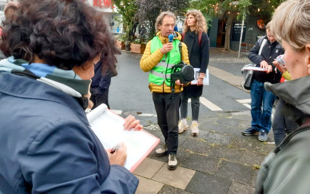 Eine Gruppe steht an einer Straßenecke, eine Frau macht Notizen, der Spaziergangsforscher Bertram Weisshaar informiert die Gruppe, hinter ihm steht die Kölner Fußverkehrsbeauftragte Britta Buch.