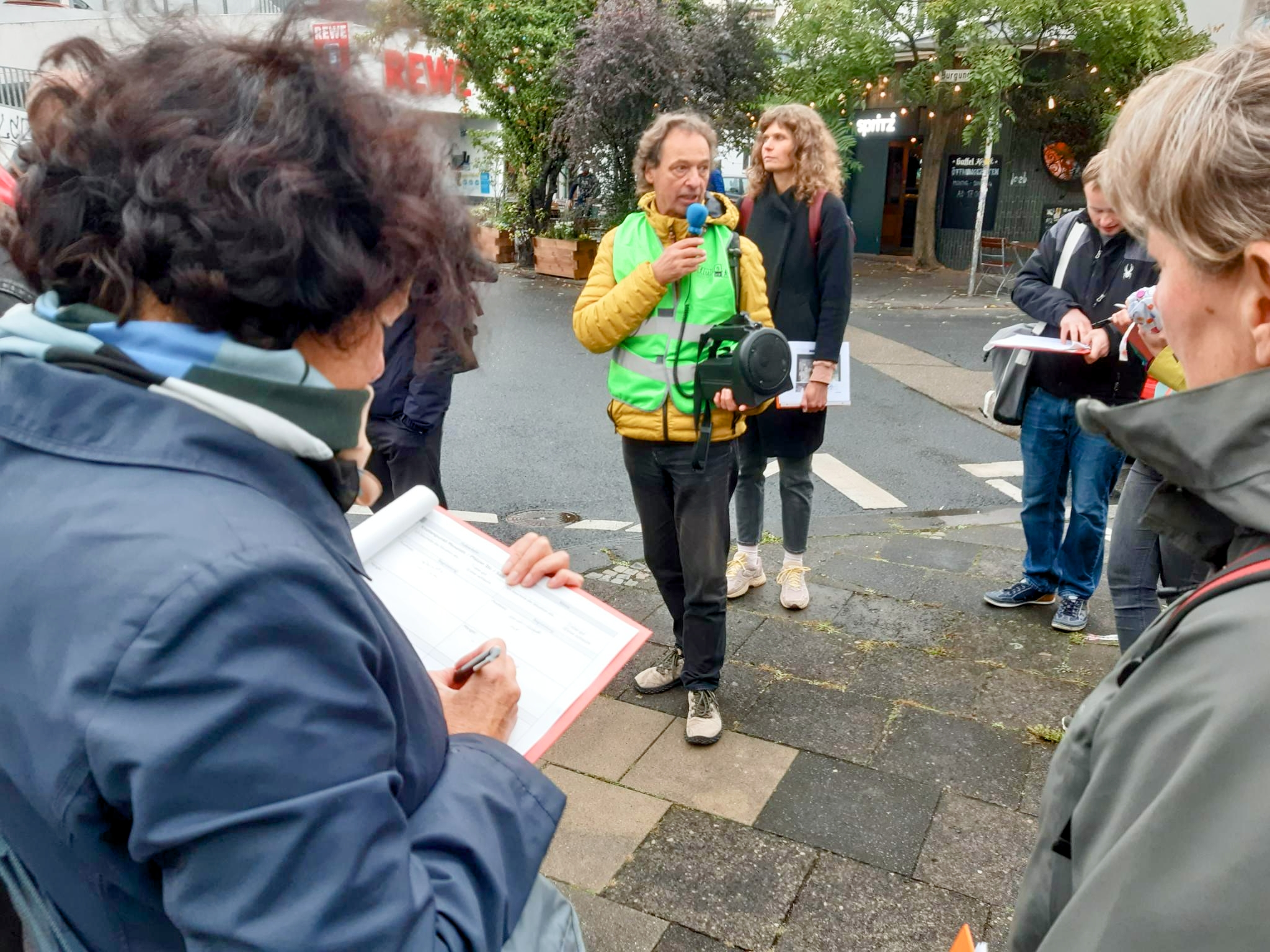 Eine Gruppe steht an einer Straßenecke, eine Frau macht Notizen, der Spaziergangsforscher Bertram Weisshaar informiert die Gruppe, hinter ihm steht die Kölner Fußverkehrsbeauftragte Britta Buch.