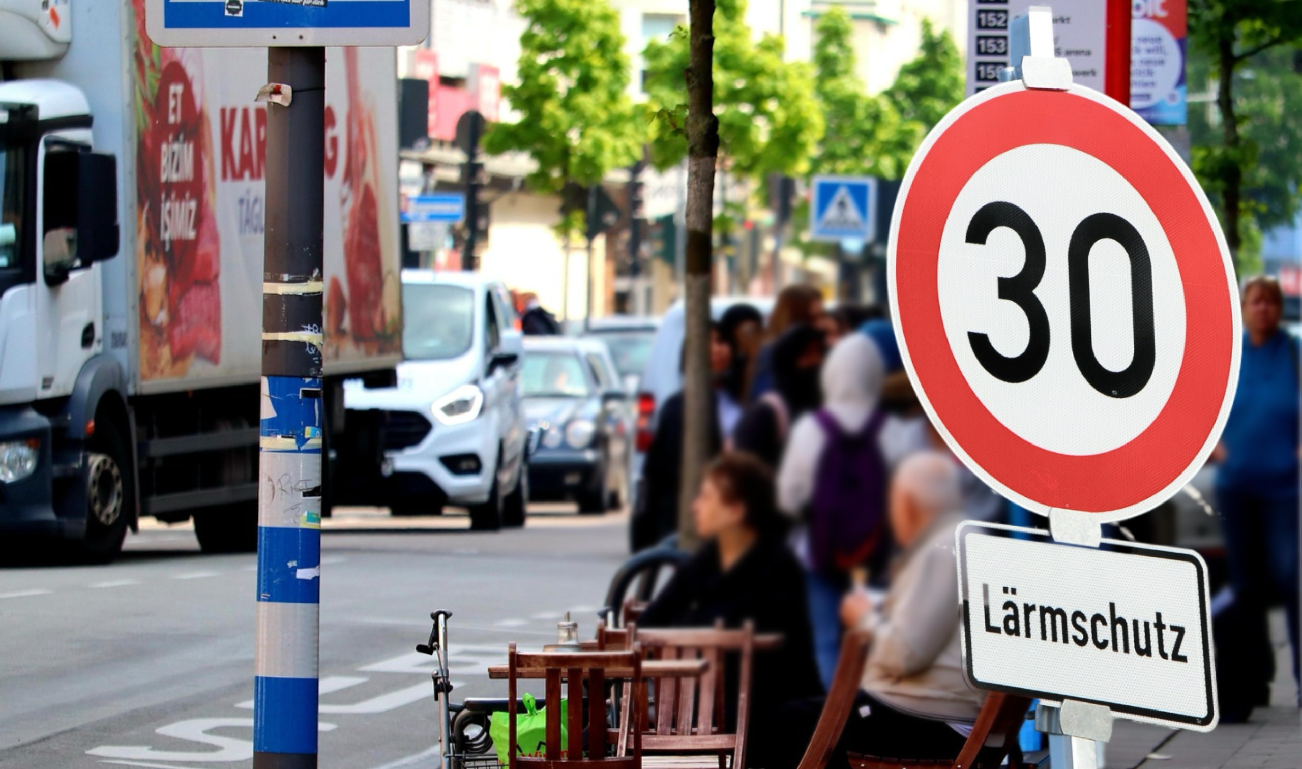 Ein Tempo-30-Schild mit dem Zusatz Lärmschutz an einer Straße, an der Menschen auf Cafestühlen sitzen, auf der anderen Seite viele Autos im Stau