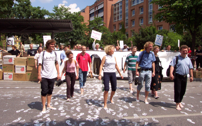 Demo: Menschen laufen barfuss und hinterlassen weiße Fußspuren auf dem Asphalt, Demoschilder und Protestbanner im Hintergund.