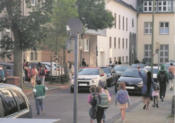 Kinder queren vor der Schule eine befahrene Straße.